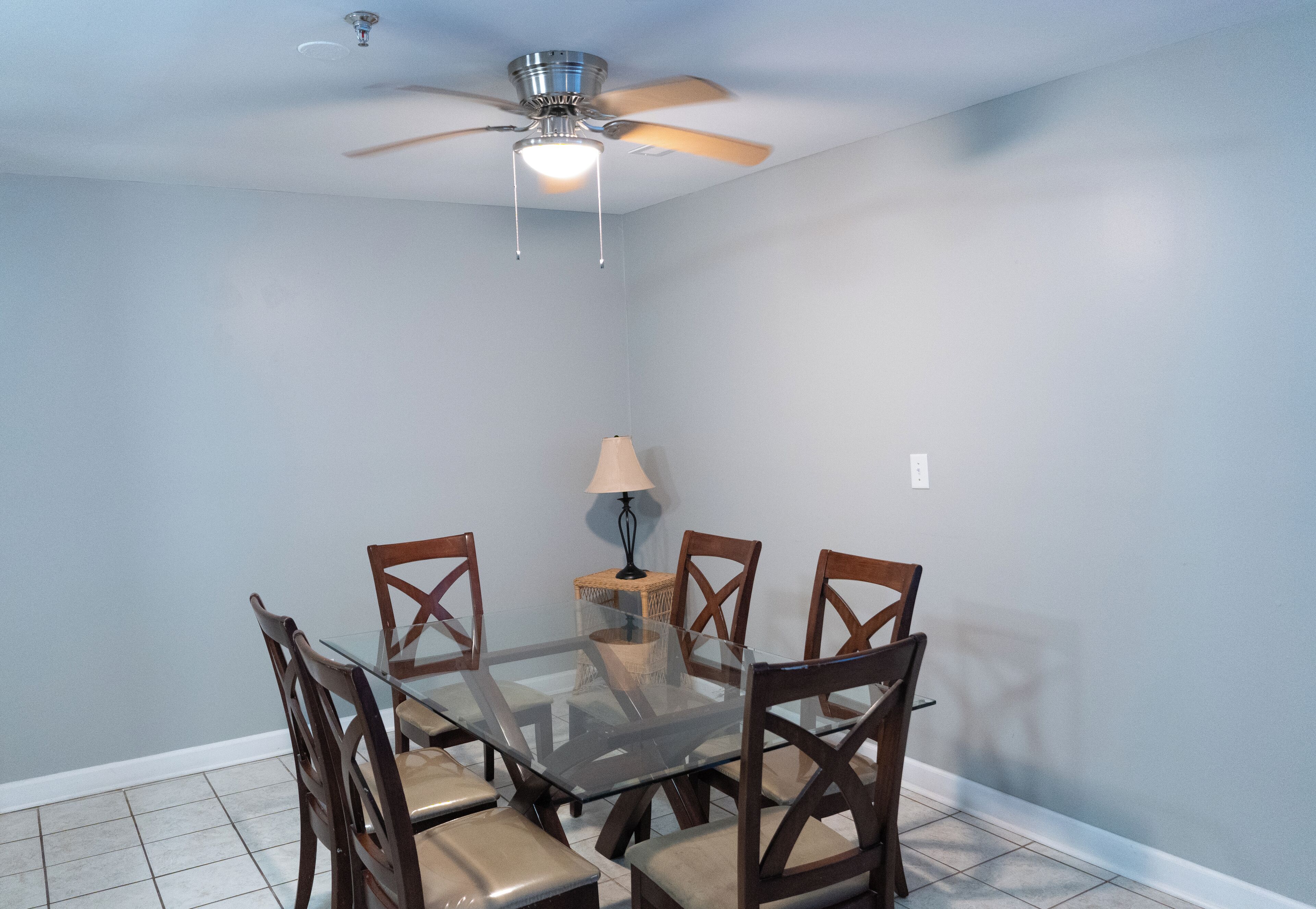 A dining area with a glass table and wooden chairs, a ceiling fan, and a lamp on a small table.