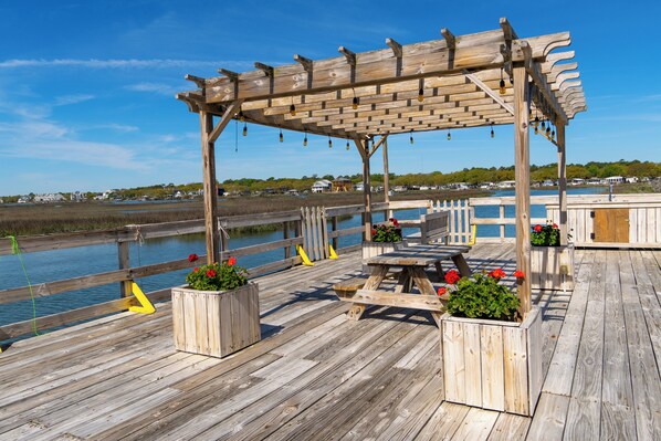 A wooden deck with a pergola, picnic tables, and planters with flowers.