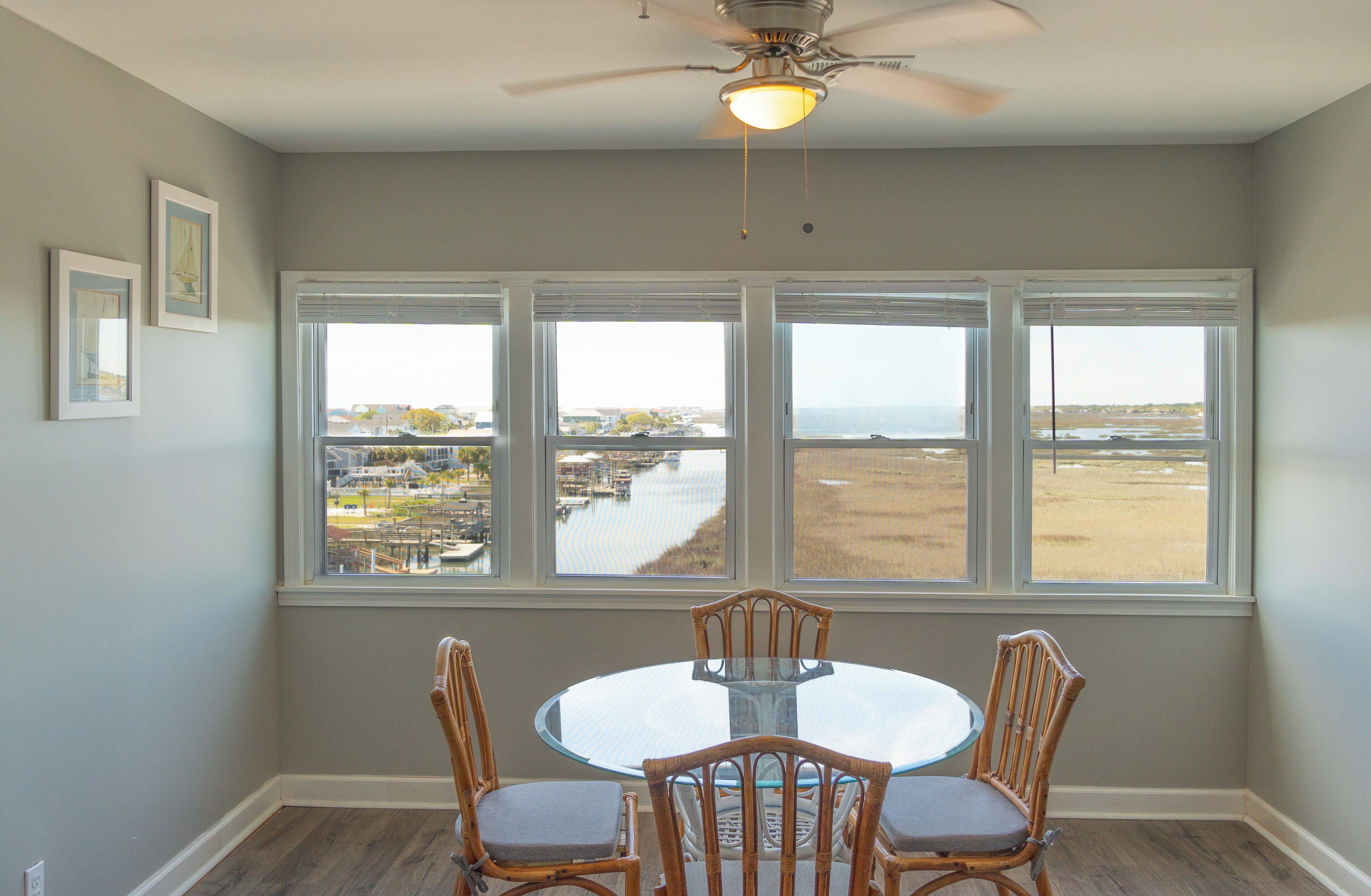 A dining area with a round glass table and four wicker chairs, large windows with a view of a river and buildings, and a ceiling fan.