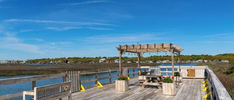 A wooden pier with a bench and a gazebo, overlooking a body of water with houses in the background.
