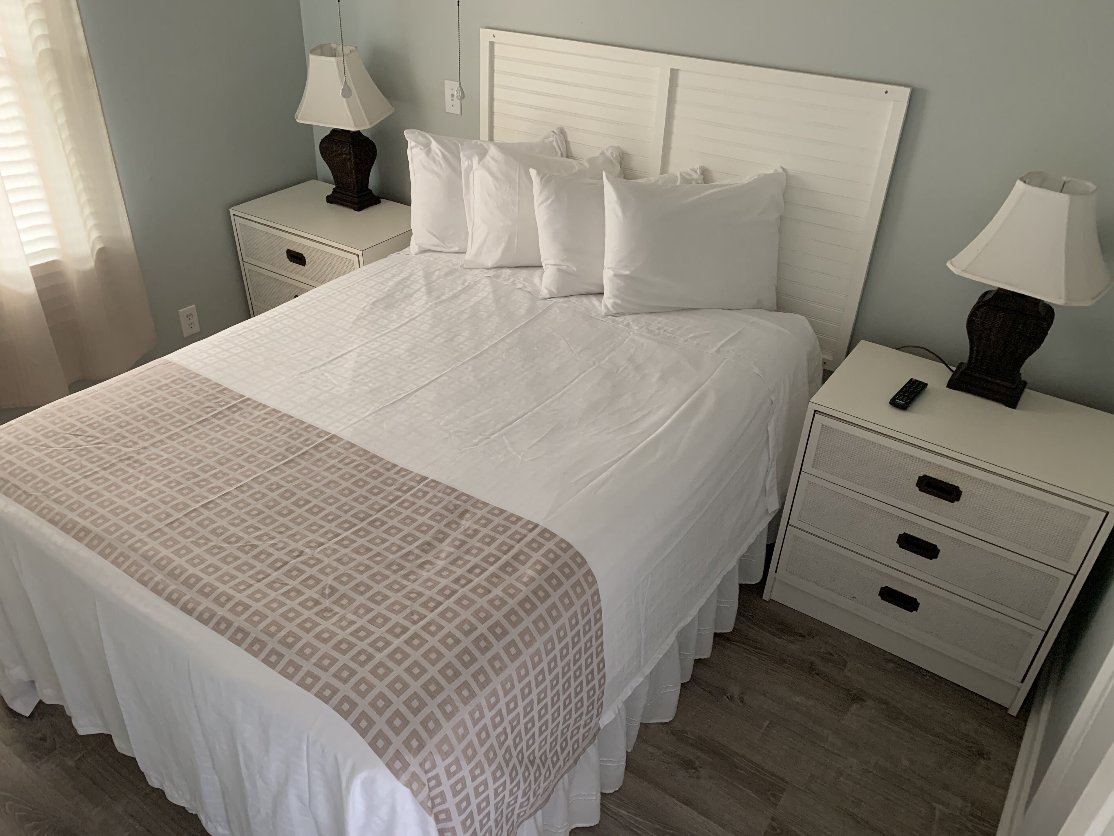 A neatly made bed with a patterned bedspread, white pillows, and a matching throw. There are two bedside tables with lamps, one of which has a remote control on it. The room has light-colored walls and wooden flooring.