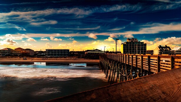 A wooden pier extending into calm waters with a sunset in the background and buildings on the shore. - Seawatch Inn at the Landing (Murrells Inlet)