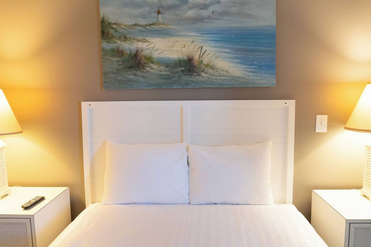 Una cama bien hecha con sábanas y almohadas blancas, flanqueada por dos mesitas de noche con lámparas, y un cuadro con una escena costera sobre la cama.