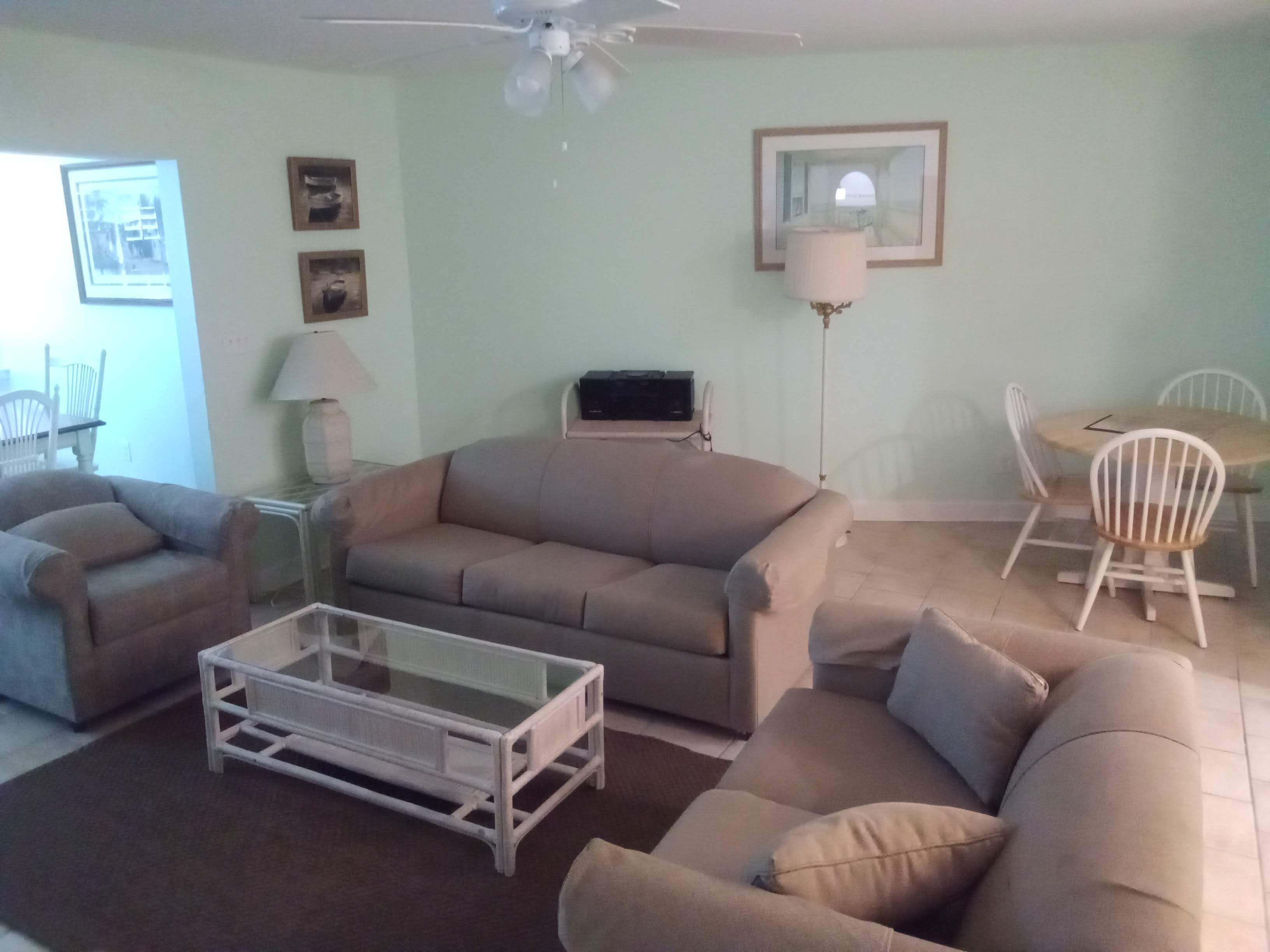 A living room with a beige sofa, a glass coffee table, a dining table with chairs, a ceiling fan, and framed pictures on the wall.