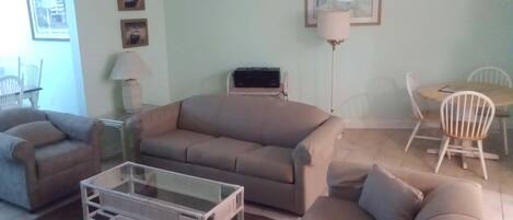 A living room with a beige sofa, a glass coffee table, a dining table with chairs, a ceiling fan, and framed pictures on the wall.