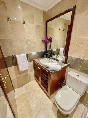 A modern bathroom with a marble floor, a wooden vanity with a sink and mirror, a toilet, and a shower area.