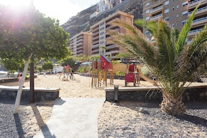 Une aire de jeux au bord de la plage, avec des balançoires et un toboggan, entourée de palmiers et avec un bâtiment visible au loin.