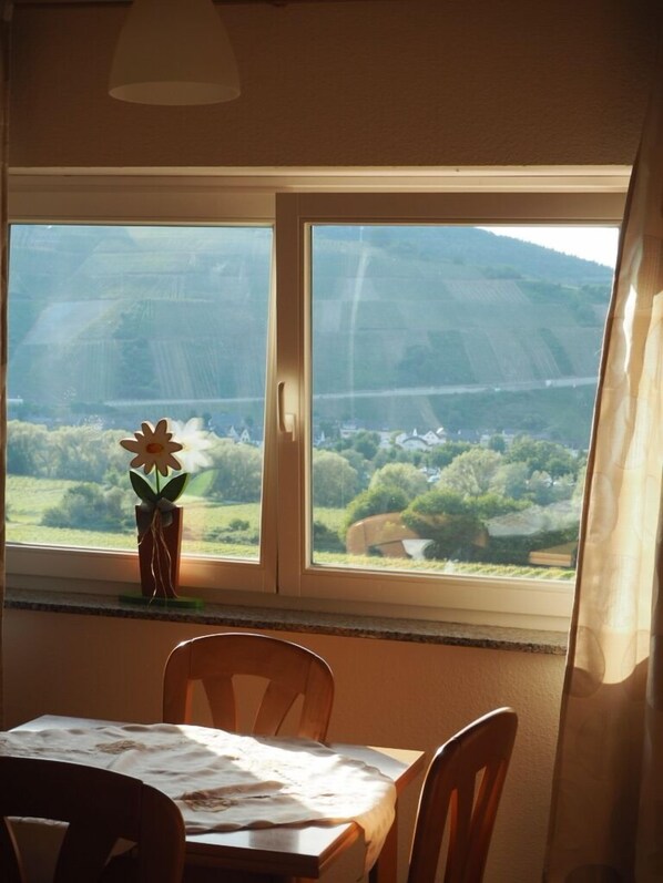 A dining area with a table and chairs, a window with a view of a hilly landscape, and a vase with a flower on the windowsill. - Weingut & Gästehaus Andreas Erz (Brauneberg)