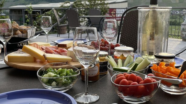 An outdoor dining setup with a variety of cheeses, meats, fruits, and vegetables on a table, with a scenic view of hills in the background. - Weingut & Gästehaus Andreas Erz (Brauneberg)
