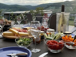 An outdoor dining setup with a variety of cheeses, meats, fruits, and vegetables on a table, with a scenic view of hills in the background. - Weingut & Gästehaus Andreas Erz (Brauneberg)