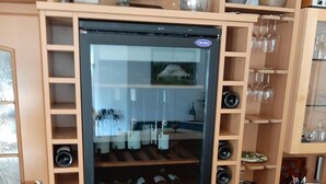 A wine refrigerator with multiple shelves for wine bottles, a glass door, and a wooden cabinet with shelves above it holding wine glasses and bottles. - Weingut & Gästehaus Andreas Erz (Brauneberg)