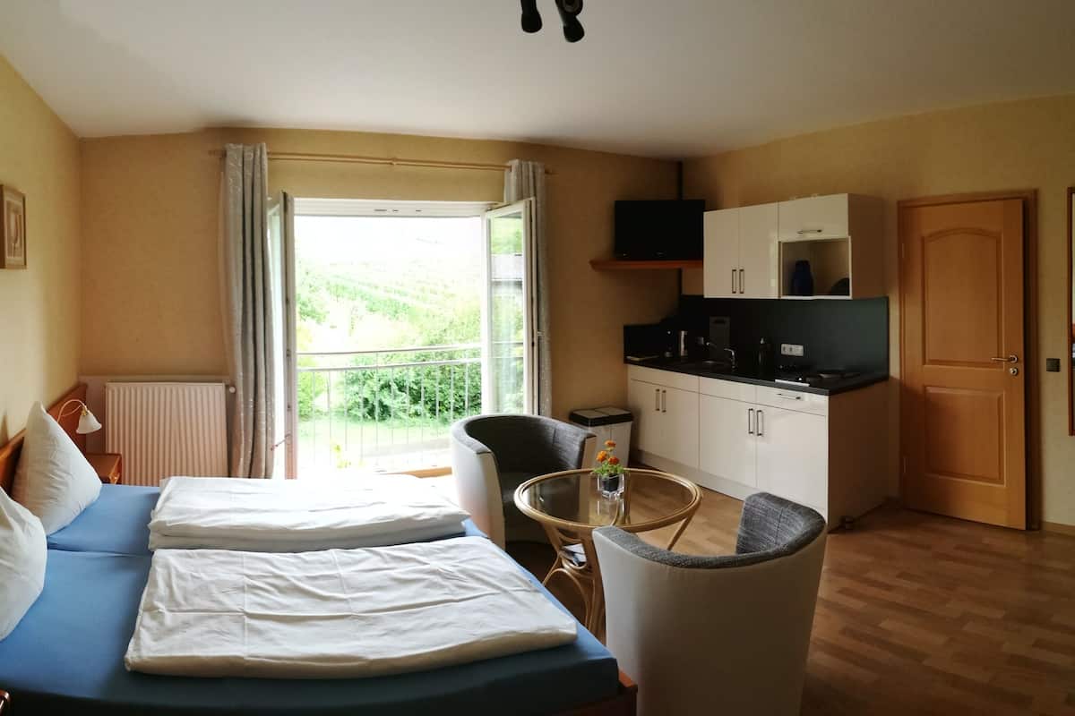 Et værelse med to senge, et lille bord, en stol, et tekøkken og et vindue med udsigt over grønne omgivelser.