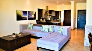 A modern living room with a sectional sofa, a coffee table, and a kitchen in the background. - BEAUTIFUL CONDO 2BD 3 MIN FROM BEACH AND DOWNTOWN (Cabo San Lucas)