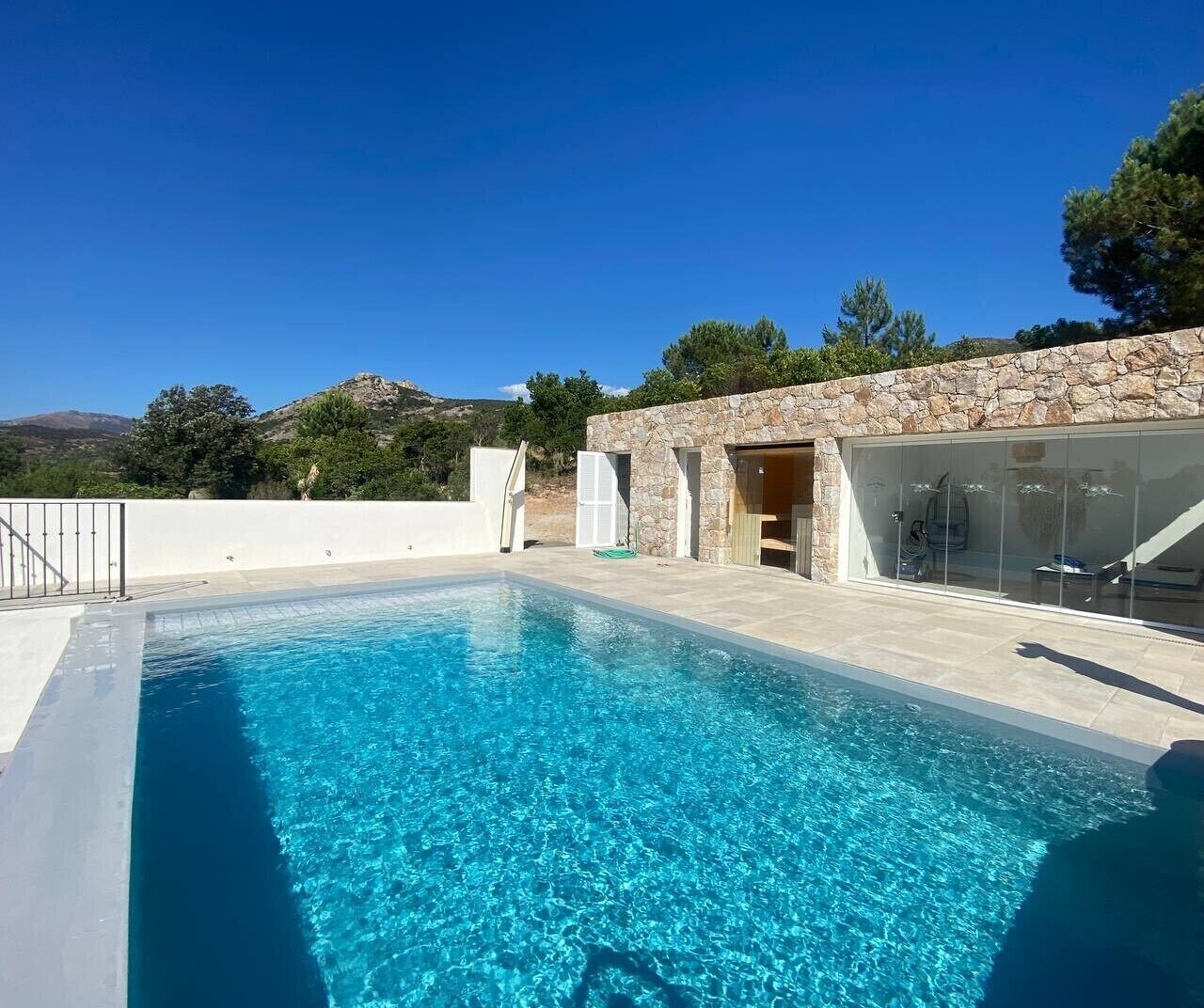 Una piscina con pared de piedra y puertas de vidrio, rodeada por un cerco blanco y árboles.
