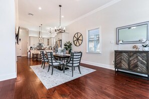 A spacious dining area with a wooden floor, a round table, and chairs. There is a chandelier above the table and a decorative wheel on the wall.