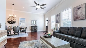 A spacious living room with a black leather sofa, a dining area with a chandelier, and artwork on the walls. - Lux Brand New! Beautiful House w/ Pool & Hot Tub (New Orleans)
