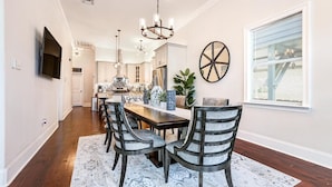A dining area with a wooden table, six chairs, a chandelier, and a large window with blinds. - Lux Brand New! Beautiful House w/ Pool & Hot Tub (New Orleans)