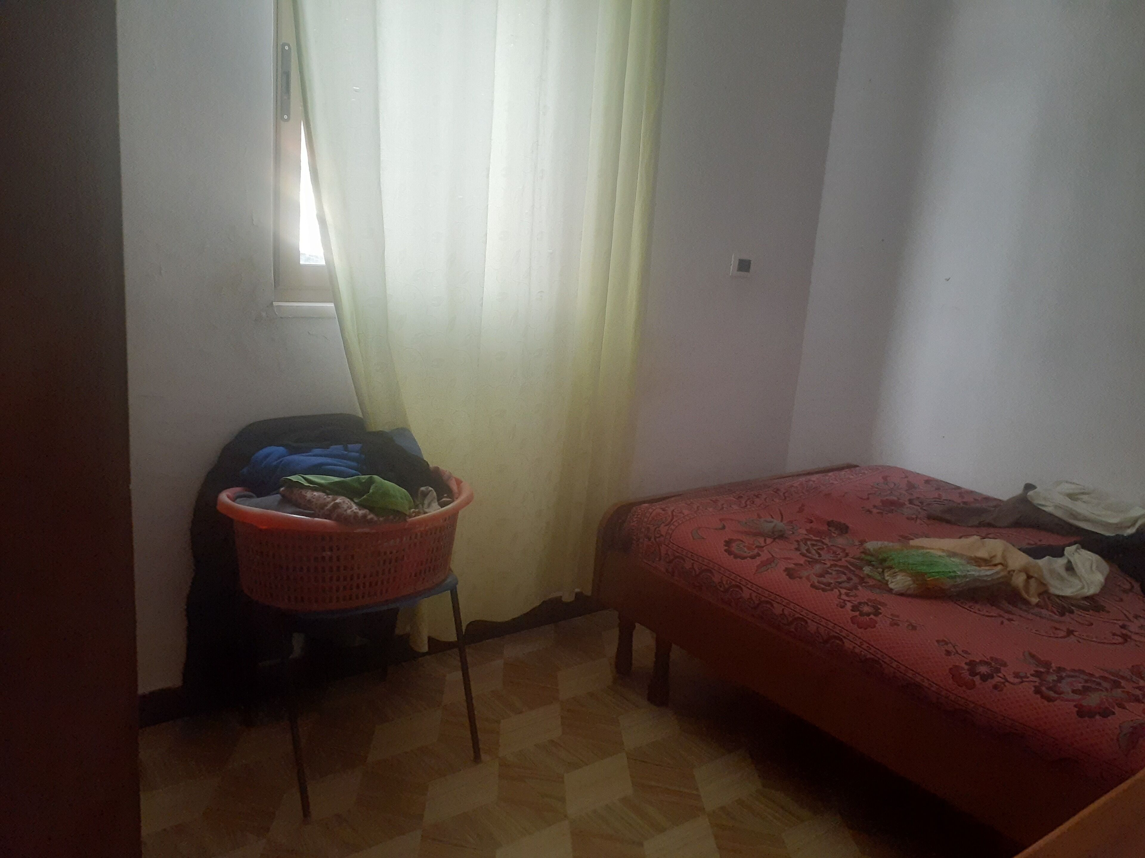A small room with a bed, a chair, and a basket of clothes.