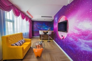 Ein Zimmer mit gelbem Sofa, einem Glastisch, einem wandmontierten Fernseher und einem Wandgemälde zum Thema Galaxie.