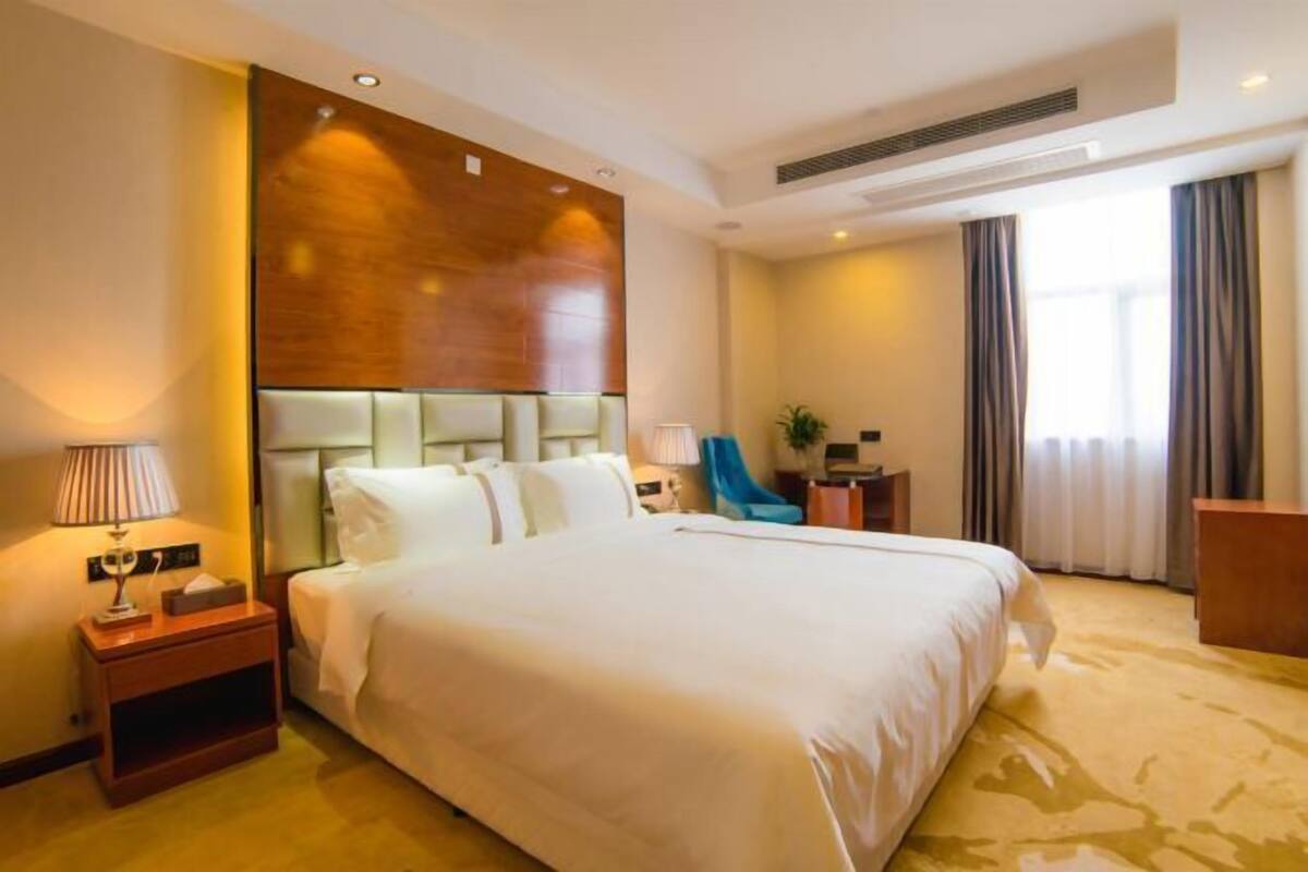 A hotel room with a large bed, bedside table, lamp, chair, and a desk with a plant.
