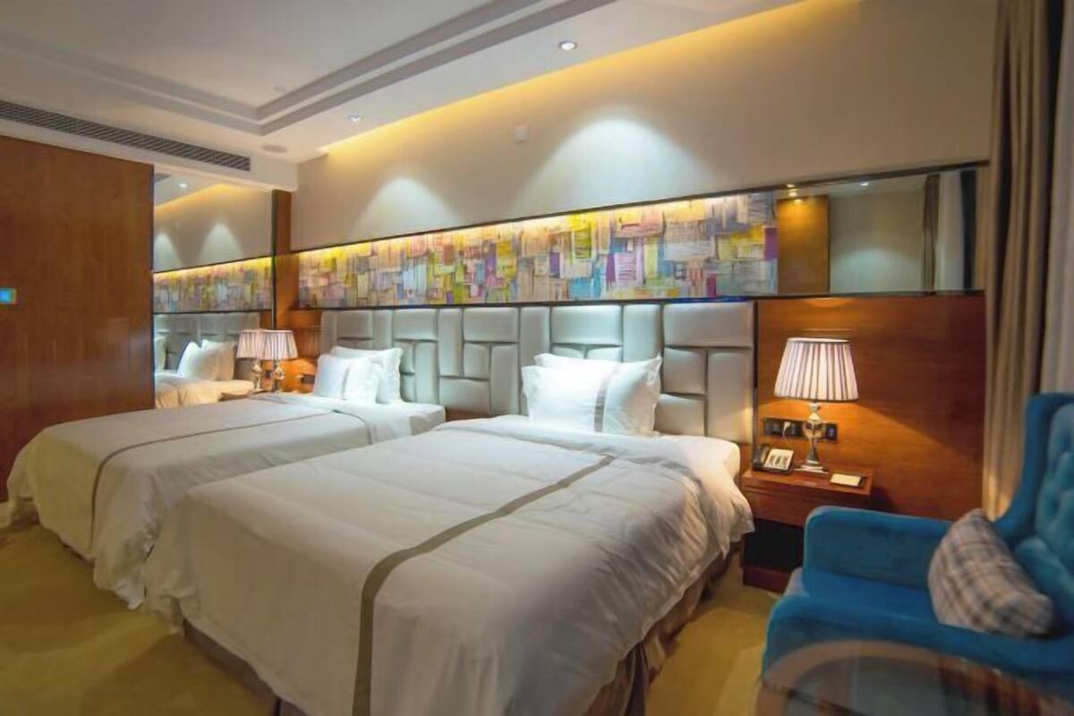 A hotel room with two beds, a blue armchair, a nightstand with a lamp, and a wall with a colorful abstract art piece.