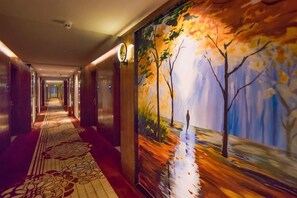 A hotel corridor with a long hallway, carpeted floor, and a mural on the wall depicting a tree and a reflective surface.