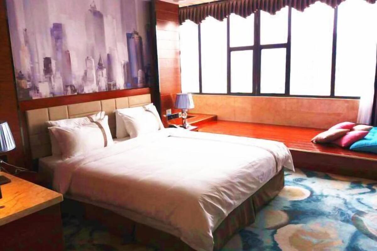 A hotel room with a large bed, a nightstand, a lamp, a window with a view, and a painting on the wall.