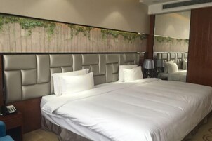A hotel room with a large bed, a headboard with a textured design, a bedside table with a lamp, and a wall-mounted mirror.