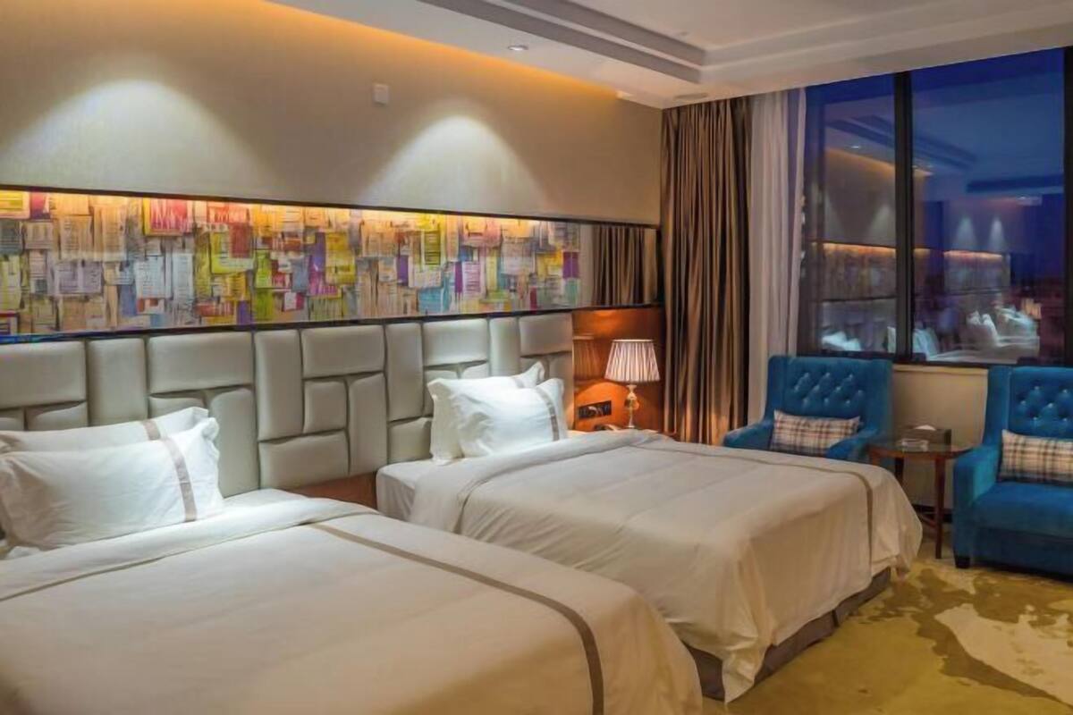 A hotel room with two beds, a large abstract wall art piece, a blue armchair, and a view of the city at night.