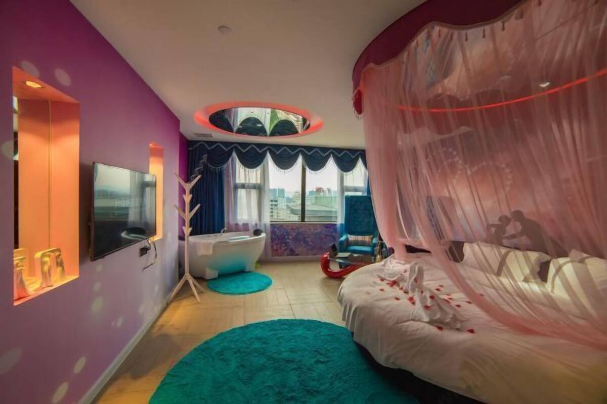 A bedroom with a bed, a bathtub, a TV, and a large window.