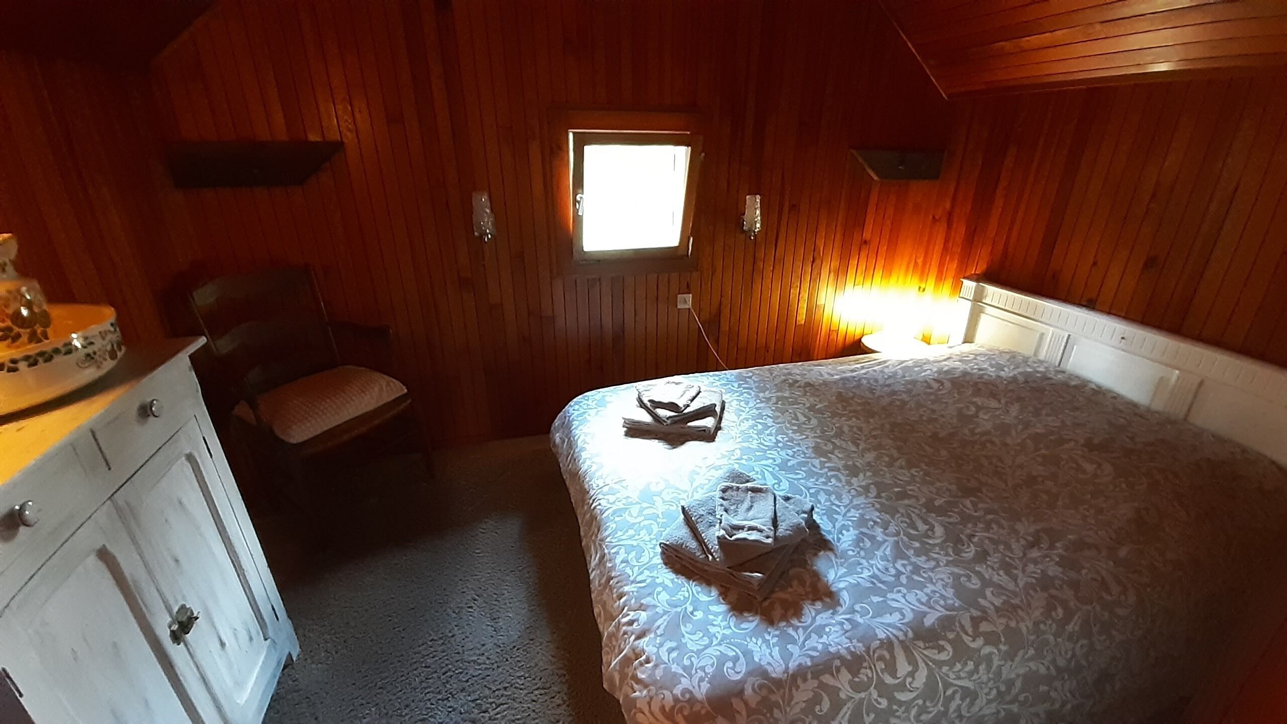 Een slaapkamer met een houten plafond, een enkel raam, een bed met een beddeken met patroon, een stoel en een dressoir.