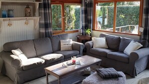 Uma sala de estar aconchegante com dois sofás, uma mesa de centro e um tapete pequeno. Há prateleiras com objetos decorativos e uma janela com cortinas.