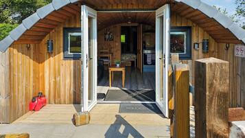 A wooden cabin with a sloped roof, a welcome mat at the entrance, a red gas canister, and a small table with a chair.