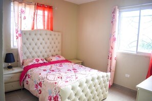 Une chambre à coucher avec une tête de lit capitonnée, une literie à motifs floraux, une lampe de chevet et une fenêtre avec des rideaux.