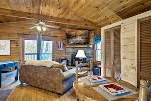 Interior acogedor de una cabaña con techo de madera, chimenea, sofá, mesa de comedor con individuales y televisor montado en la pared.