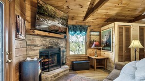 Ein gemütliches Blockhaus-Interieur mit Kamin aus Naturstein, Holzwänden, einer Couch und einem Fenster mit Blick auf Bäume.