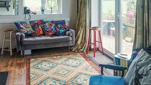 A cozy living room with a grey sofa, colorful pillows, a patterned rug, a wooden stool, and a large window with a view of the outdoors.