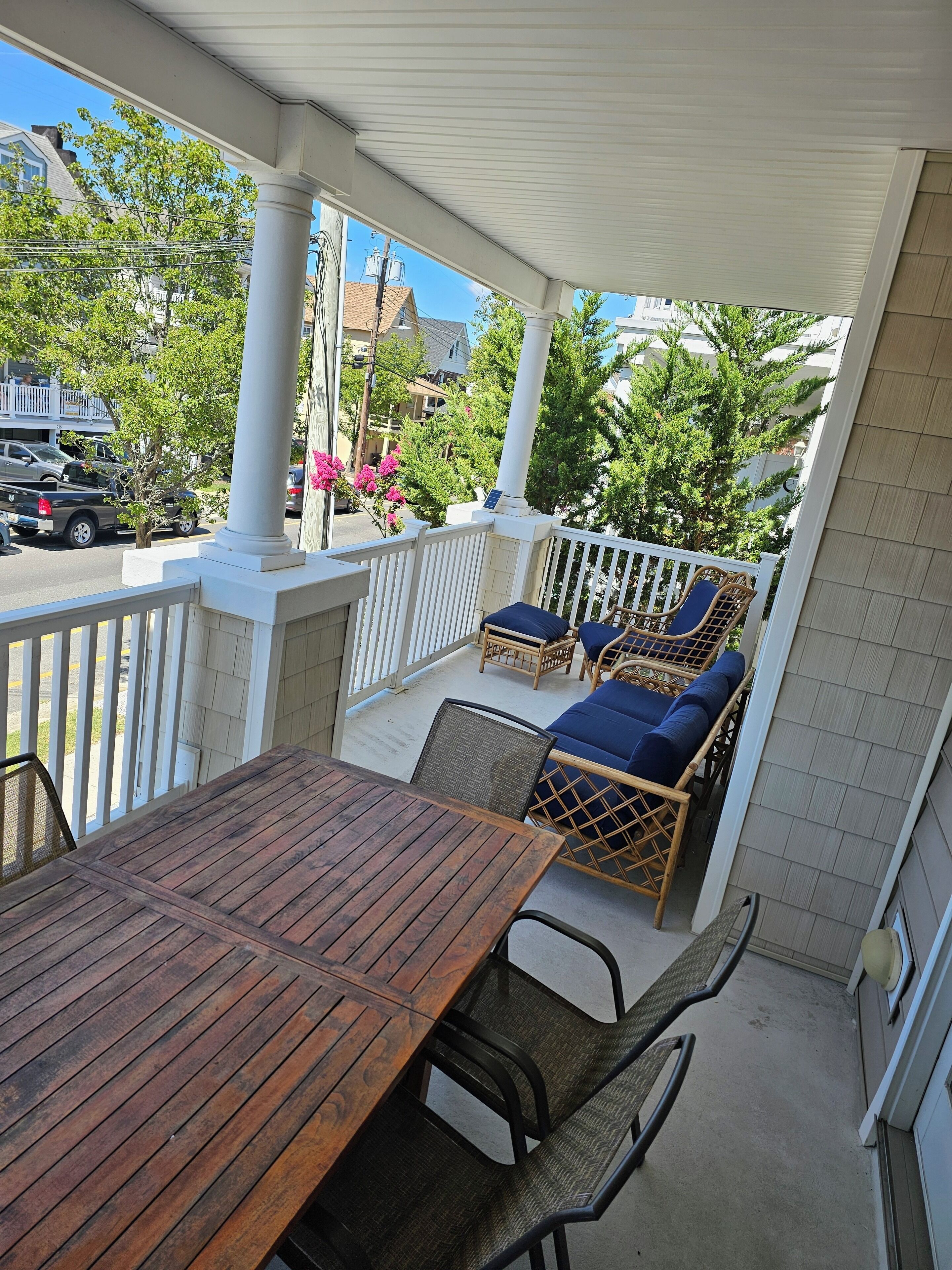 Eine überdachte Terrasse im Freien mit Holztisch und Korbstühlen, einem weißen Geländer und Blick auf die Straße und die Gebäude.