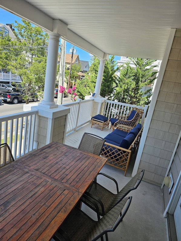 A covered outdoor patio with a wooden table and wicker chairs, a white railing, and a view of the street and buildings. - 3BR, 2BA Wildwood condo, 2 blocks to beach, parking, linens, 5 queen beds. (Wildwood)