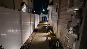 Un étroit sentier bordé de clôtures blanches et de coffrets de raccordement, qui mène à un bâtiment dont l’entrée est illuminée de lumière bleue.