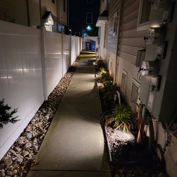 A narrow pathway flanked by white fences and utility boxes, leading to a building with a blue-lit entrance. - 3BR, 2BA Wildwood condo, 2 blocks to beach, parking, linens, 5 queen beds. (Wildwood)