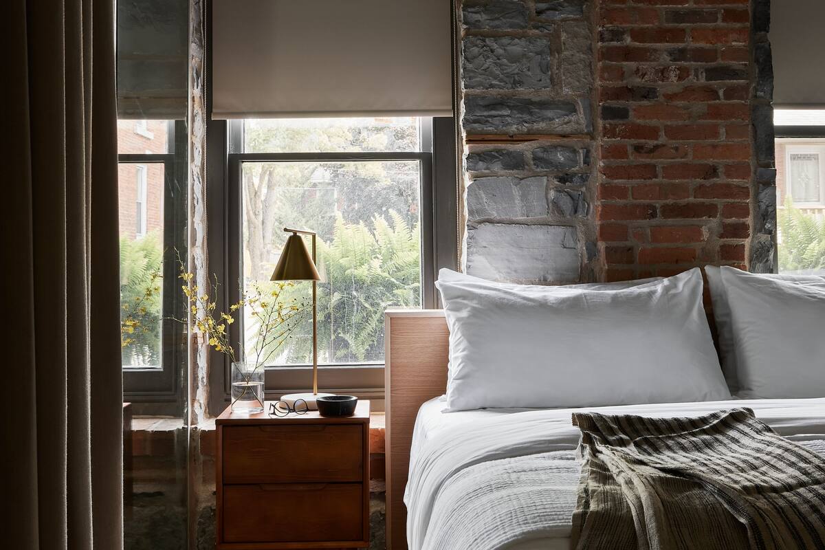 Un dormitorio con una cama grande, una mesita de noche, una ventana con vistas a zonas verdes y una pared de ladrillo.