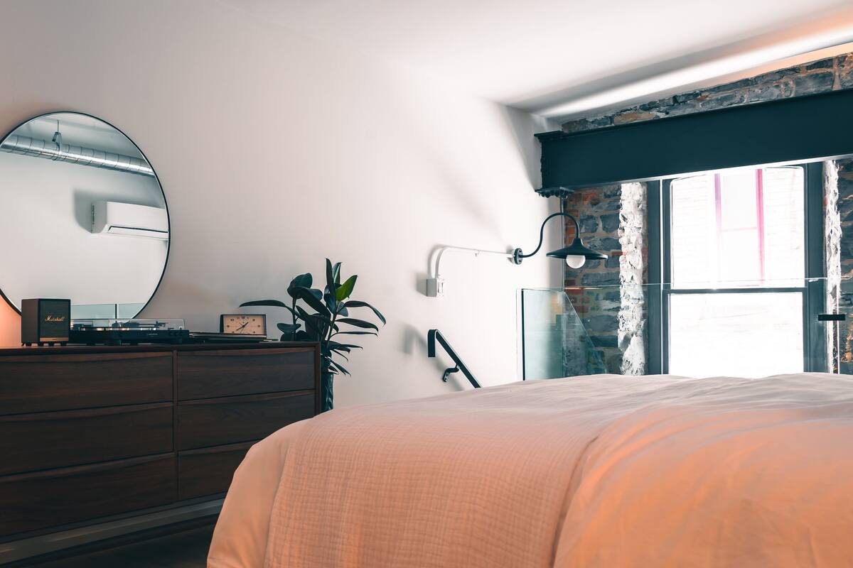 Un dormitorio con cama, cómoda, espejo, planta y ventana con vista a un muro de piedra.