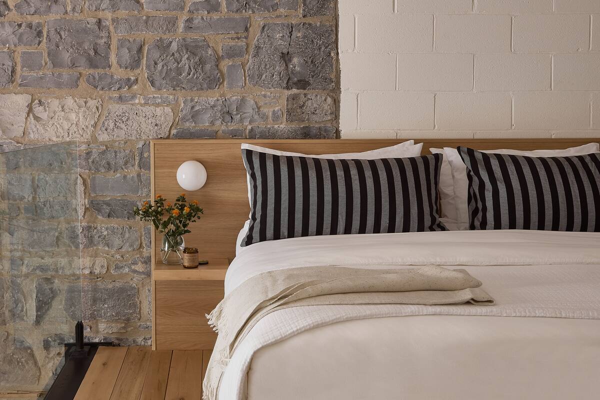Un dormitorio con pared de piedra, estructura de cama de madera y cabecera blanca.