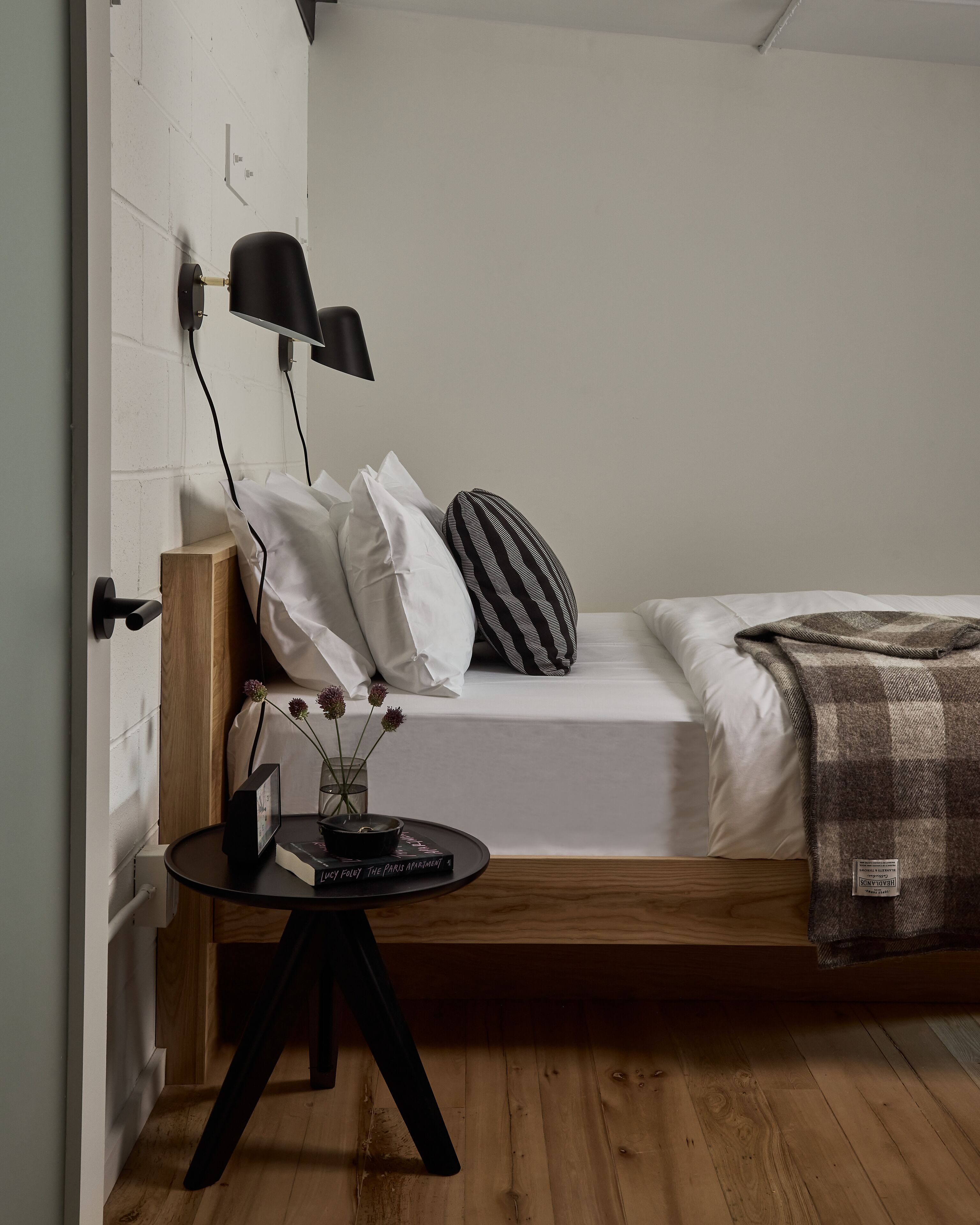 Une chambre à coucher de taille modeste, comprenant un lit, une table de chevet, une lampe et une petite table sur laquelle sont posés un livre et des fleurs.