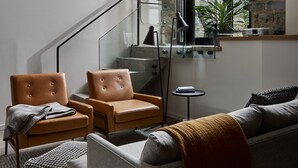 A modern living room with a staircase, leather armchairs, a sofa, and a large window with a view of a brick building.