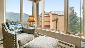 Un coin salon avec un fauteuil et un pouf à motifs, une table avec une lampe et une grande fenêtre donnant sur les montagnes.
