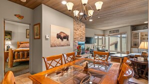 Un espace repas comprenant une table à plateau de verre et des chaises en bois, un mur décoratif en pierre, ainsi qu’un plafond orné de poutres en bois et de lustres.