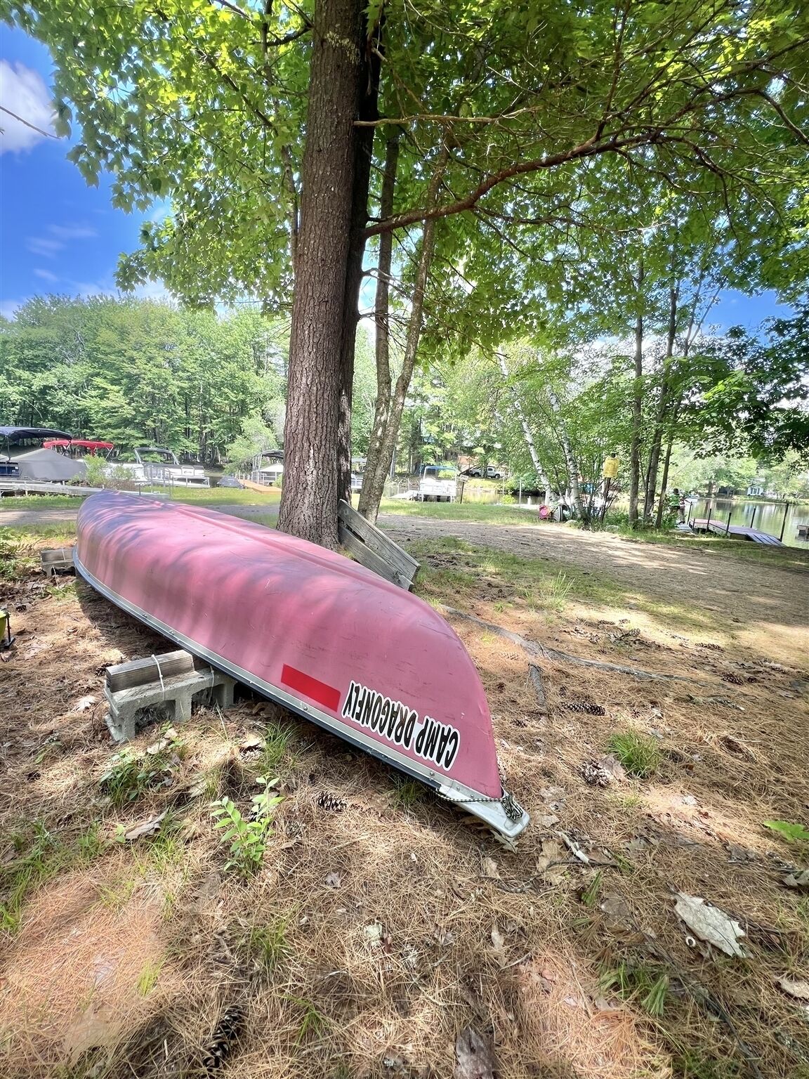 A canoe with "CAMP DRAGONCET" written on it, resting on a concrete block under a tree in a wooded area.