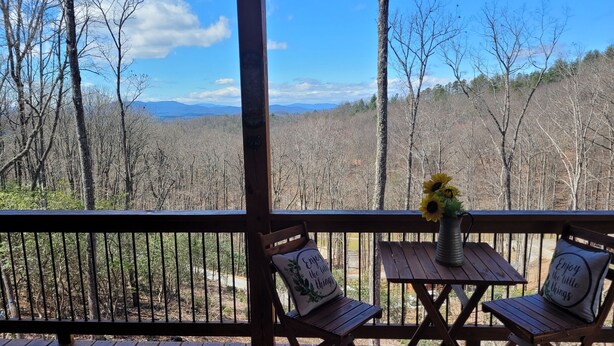 A wooden deck with two wooden chairs and a small table, each with a cushion. A vase with sunflowers is on the table. The deck overlooks a forested area with mountains in the distance. - Mountain View Cabin,15 Min to Casino, Family Friendly (murphy)